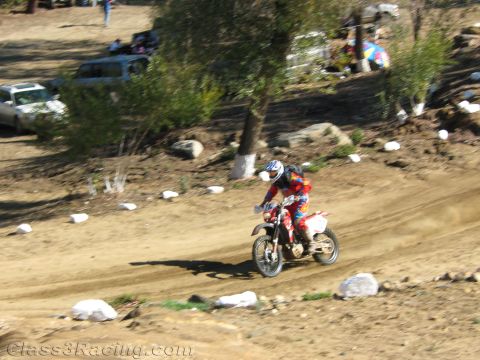 1990 Ford Bronco - dirt-bike rider rides a sweeping dirt track lined with white marker rocks, with parked vehicles and spectators in the distance.