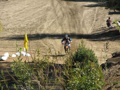 1990 Ford Bronco - dirt-bike rider kicks up dust riding toward the camera on a hillside track, viewed over green brush and a yellow course flag.