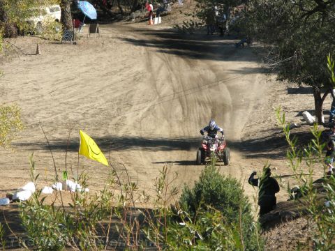 1990 Ford Bronco - rider on a quad ATV speeds up a dusty hill trail past a yellow smiley-face flag with spectators nearby.