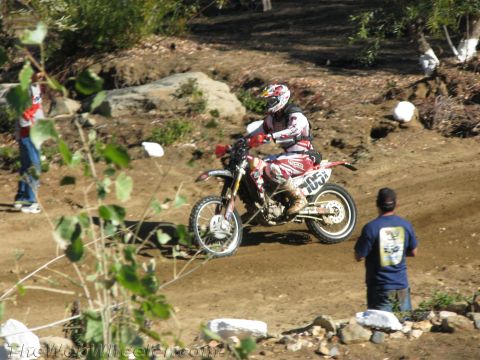 1990 Ford Bronco - dirt bike rider in red and white gear, bike numbered 1052, rides a dirt course as a man in a blue shirt watches.