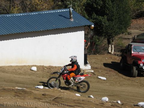 1990 Ford Bronco - rider on dirt bike number 310 passes a white building with a blue metal roof, with a red Jeep parked nearby.