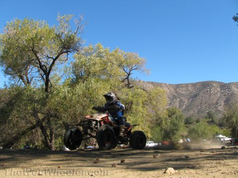 1990 Ford Bronco - rider catches air on a red and black quad ATV over open dirt with a tree, mountains and clear blue sky behind.
