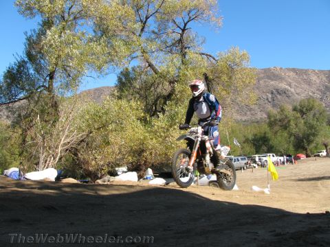 1990 Ford Bronco - dirt bike rider in blue and white gear rides on open dirt near a yellow flag, trees, parked trucks and mountains.