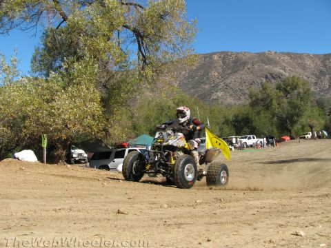1990 Ford Bronco - rider on a yellow and black quad ATV kicks up dust racing past a yellow flag with mountains in the background.