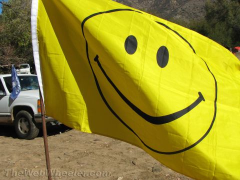 1990 Ford Bronco - large yellow smiley-face flag waves on a pole beside a white pickup truck with mountains behind.