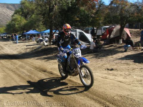 1990 Ford Bronco - rider on blue dirt bike number 304 rides a dirt track with tents and spectators in the background.