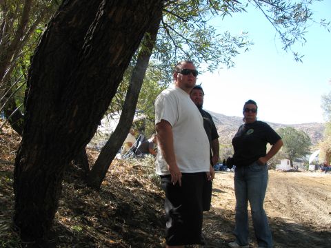 1990 Ford Bronco - Three people, two men and a woman, stand under a tree on a dirt lot at an outdoor off-road gathering.