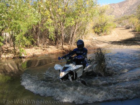 1990 Ford Bronco - rider on a white dirt bike crosses a shallow creek, throwing up water spray with trees and a hillside behind.