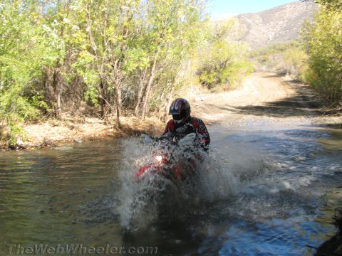 1990 Ford Bronco - rider on a red dirt bike charges through a creek crossing, sending up a large spray of water on a trail.