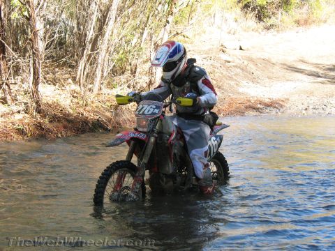 1990 Ford Bronco - rider on dirt bike number 176x pauses in a shallow creek crossing with brush and a dirt road behind.