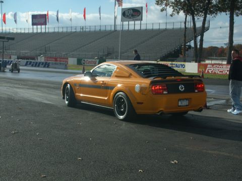 2001 Mercury Grand Marquis - A yellow-orange Ford Mustang is parked on a drag strip with a grandstand in the background.