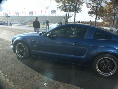 2001 Mercury Grand Marquis - A blue Ford Mustang GT is parked on the asphalt at a drag strip, with spectators and bleachers in the background.