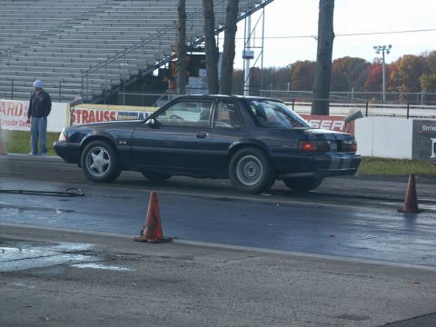 2001 Mercury Grand Marquis - A dark blue Ford Mustang hatchback, likely from the late 1980s, sits on a drag strip with racing stripes on its rear.