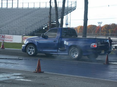 2001 Mercury Grand Marquis - A blue pickup truck is seen on a race track with smoke billowing from its rear tire.