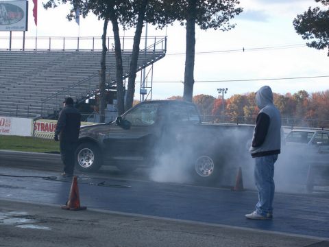 2001 Mercury Grand Marquis - A truck performs a burnout on a drag strip, creating a large cloud of smoke as onlookers watch.