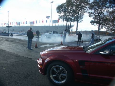 2001 Mercury Grand Marquis - A drag strip scene with a red Mustang GT in the foreground and a pickup truck performing a burnout.