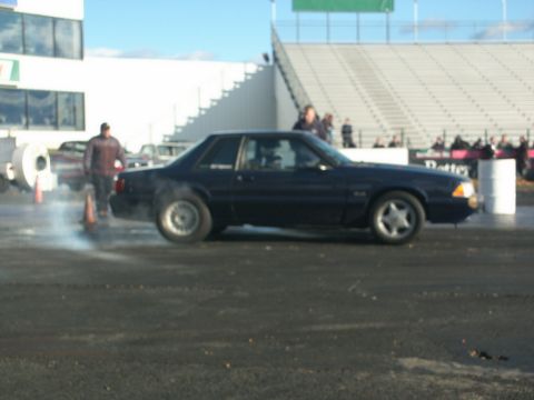 2001 Mercury Grand Marquis - A dark blue coupe performs a burnout on a drag strip, with spectators in the background.