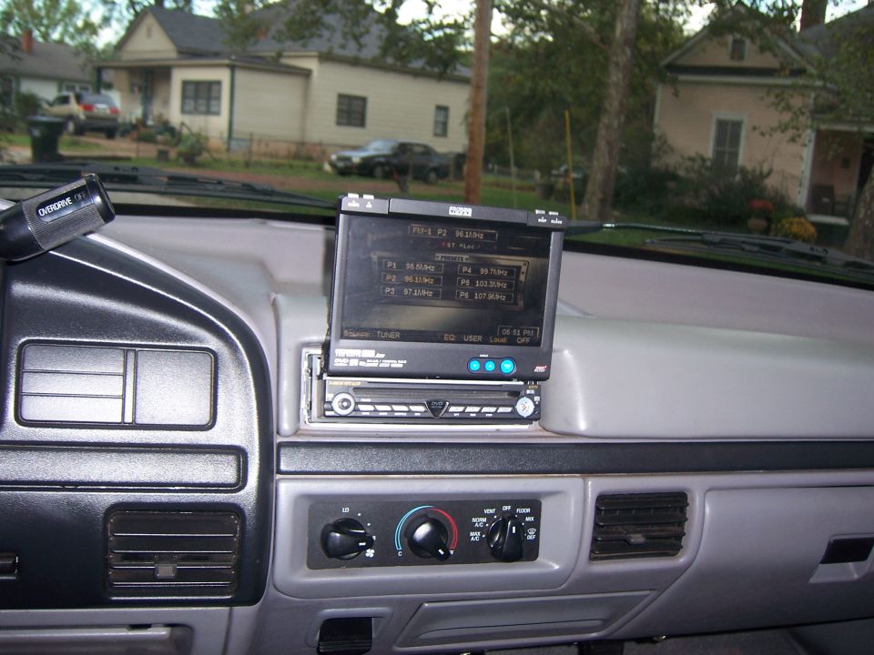 Gray dash with aftermarket flip-out DVD touchscreen radio showing FM presets, factory HVAC knobs below