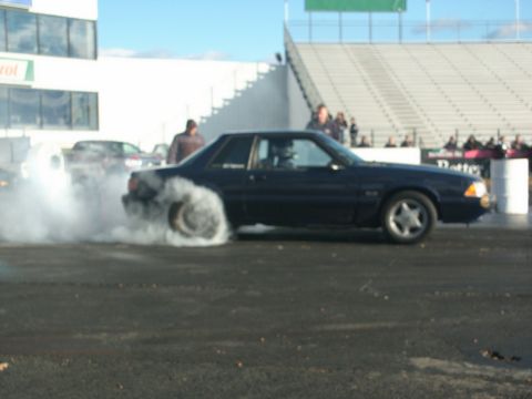 2001 Mercury Grand Marquis - A dark coupe performs a burnout on a track, with spectators in the stands and another vehicle in the background.