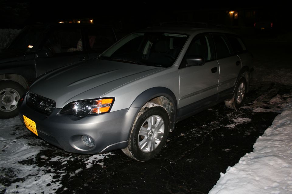 Silver Subaru Outback wagon at night, 3/4 front view, yellow plate, on snowy lot