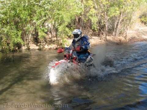 1990 Ford Bronco - rider on a red dirt bike speeds through a creek crossing, throwing up a wide spray of water amid brush.