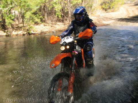 1990 Ford Bronco - rider on an orange dirt bike numbered 083, headlight on, crosses a creek splashing water with brush behind.