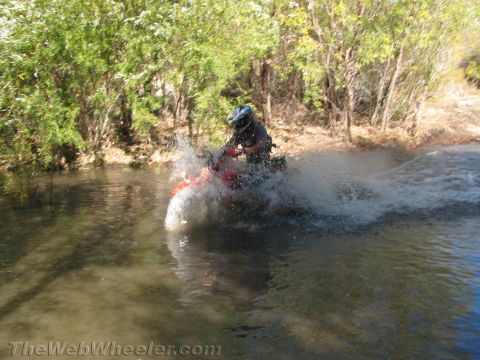 1990 Ford Bronco - red dirt bike rider is partly hidden in a large spray of water while crossing a creek lined with brush.