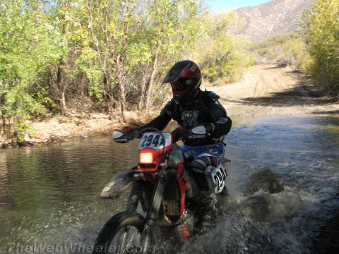 1990 Ford Bronco - rider on dirt bike number 294 splashes through a creek on a canyon trail with brush and hills behind.