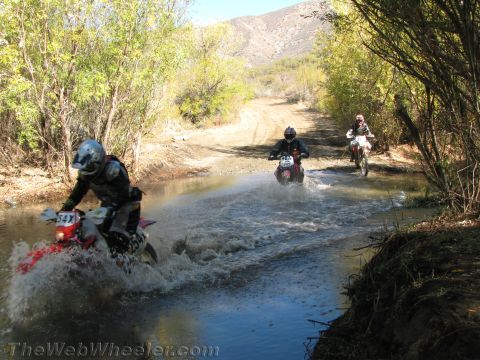 1990 Ford Bronco - Three dirt bike riders cross a creek on a dirt trail one behind another, with brush and mountains behind.