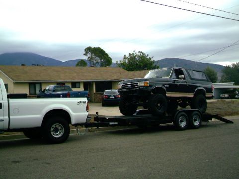 2000 Ford F-250 - A lifted black Ford Bronco is transported on a trailer, towed by a white pickup truck.