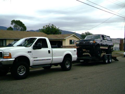 2000 Ford F-250 - A white Ford F-250 tows a lifted black Ford Bronco on a trailer down a residential street.