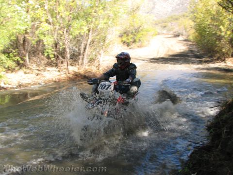 1990 Ford Bronco - rider on dirt bike number 707 charges through a creek crossing, throwing up water spray on a brush-lined trail.