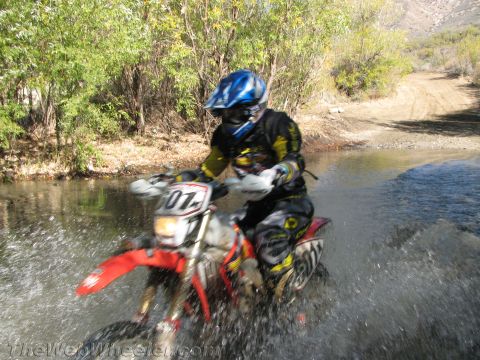 1990 Ford Bronco - rider in a blue helmet and yellow-black gear powers through a creek on a red dirt bike, spraying water.