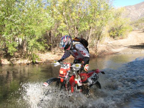 1990 Ford Bronco - helmeted rider plows a red off-road motorcycle, number 262, through a shallow river crossing, kicking up a wide spray of water.