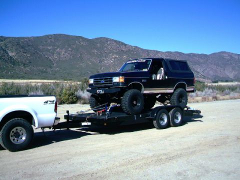 2000 Ford F-250 - White Ford F-250 tows a lifted black Ford Bronco on a trailer through a dry, mountainous landscape.