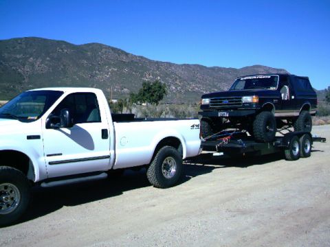 2000 Ford F-250 - A lifted Ford Bronco is transported on a trailer behind a white Ford pickup truck under a clear blue sky.