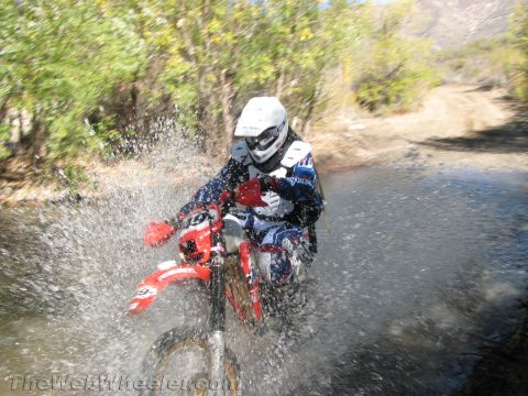 1990 Ford Bronco - rider in a white helmet and blue gear powers a dirt bike through a river crossing, throwing up a heavy curtain of spray.