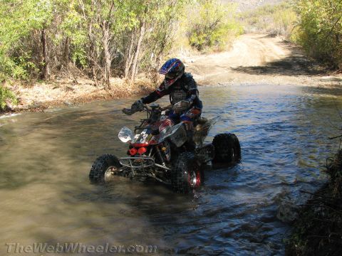 1990 Ford Bronco - rider on a sport quad ATV picks through a shallow river crossing next to a winding dirt trail in a brushy canyon.