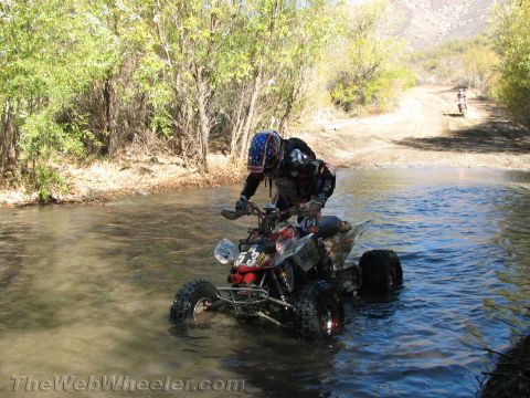 1990 Ford Bronco - rider on quad ATV number 53 wades across a river crossing while another rider approaches on the dirt road behind.