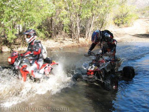 1990 Ford Bronco - Two riders push their sport quad ATVs through a river crossing side by side, one numbered 53, sending up sheets of water.
