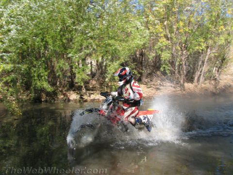 1990 Ford Bronco - rider in red and white gear blasts a dirt bike through a river crossing, throwing a tall fan of spray amid green willows.