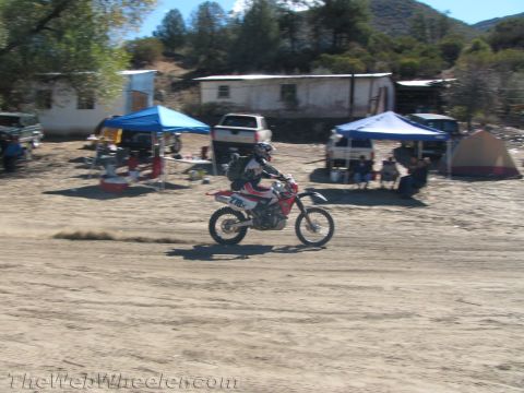 1990 Ford Bronco - dirt bike rider rolls across a sandy staging area past pop-up canopies, vehicles and an old building in a hilly setting.