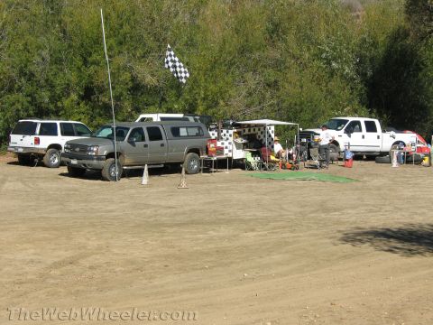 1990 Ford Bronco - row of pickups and SUVs with pop-up canopies and a checkered flag forms a pit area on a dirt clearing beside green brush.