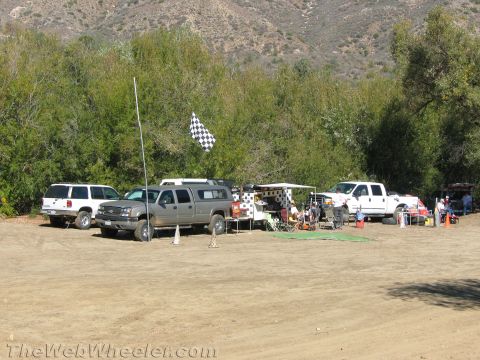 1990 Ford Bronco - Pickup trucks, an SUV and pop-up canopies line a dirt pit area flying a checkered flag below a brushy canyon hillside.