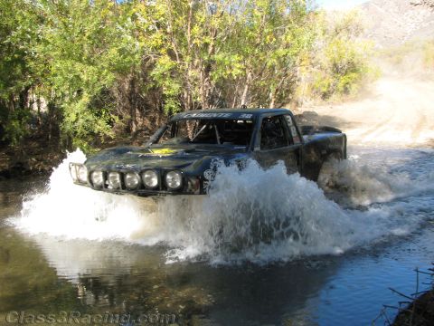 1990 Ford Bronco - tube-chassis off-road race truck, number 38 with a row of front driving lights, blasts through a river crossing in a spray of water.