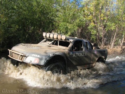 1990 Ford Bronco - mud-caked off-road race truck lettered RIVIERA with BFGoodrich and PIAA decals churns through a river crossing.