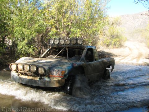 1990 Ford Bronco - weathered off-road race truck topped by a six-light roof bar wades a river crossing alongside a dusty dirt trail.