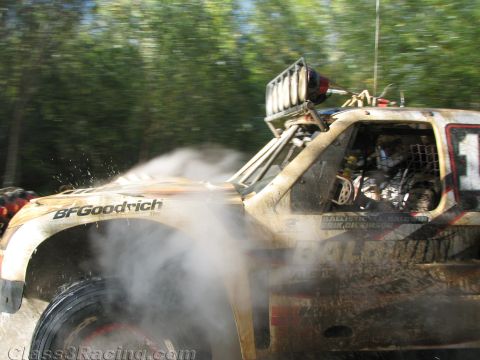 1990 Ford Bronco - mud-spattered off-road race truck with BFGoodrich livery and a roof light bar speeds past amid spray and motion blur.