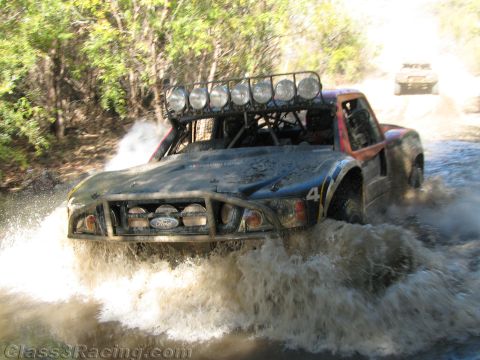 1990 Ford Bronco - off-road race truck wearing a Ford grille badge and roof light bar plows through a river crossing as another truck follows.