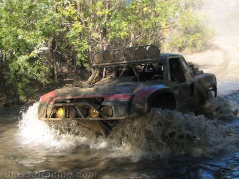 1990 Ford Bronco - dark, mud-streaked off-road race truck with glowing yellow driving lights charges through a river crossing in heavy spray.